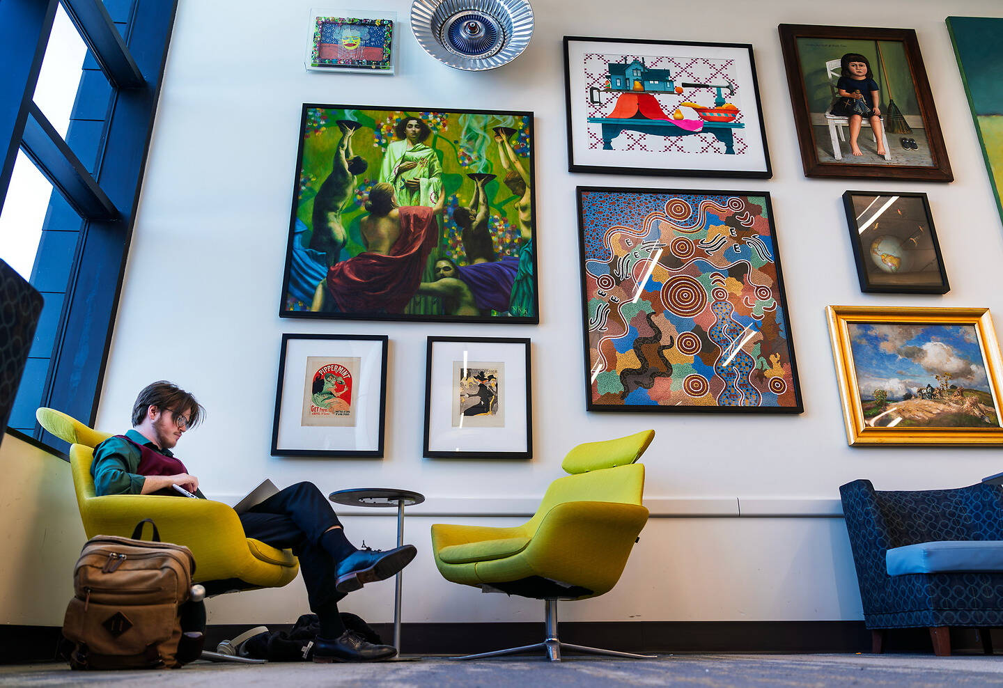 A person sits in a vibrant yellow chair, reading a book, surrounded by colorful artwork on a white wall in a modern lounge.